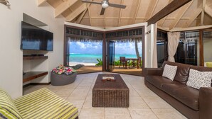 A spacious living area with a large window overlooking a beach, a comfortable sofa, a television, and a wicker coffee table. - Sea Change Villas (Rarotonga)