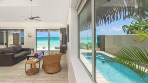 A modern living room with a large glass wall, a ceiling fan, a pool, and a view of the ocean. - Sea Change Villas (Rarotonga)