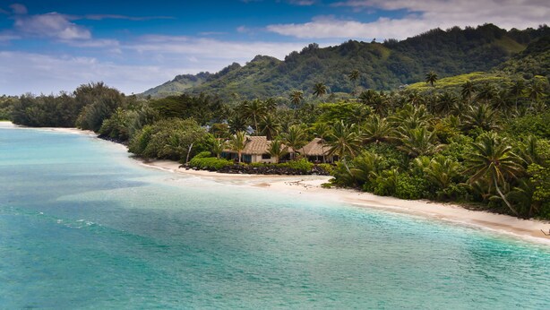 A beach with clear turquoise waters, a sandy shore, and a small cluster of thatched-roof huts surrounded by lush greenery and palm trees. - Sea Change Villas (Rarotonga)