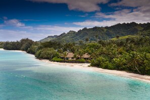 A beach with clear turquoise waters, a sandy shore, and a small cluster of thatched-roof huts surrounded by lush greenery and palm trees. - Sea Change Villas (Rarotonga)