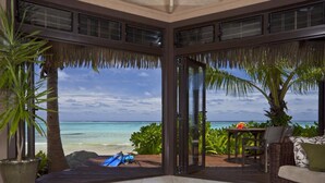 A room with a glass door leading to a beach view, a wicker sofa, a small table, and a blue chair. - Sea Change Villas (Rarotonga)
