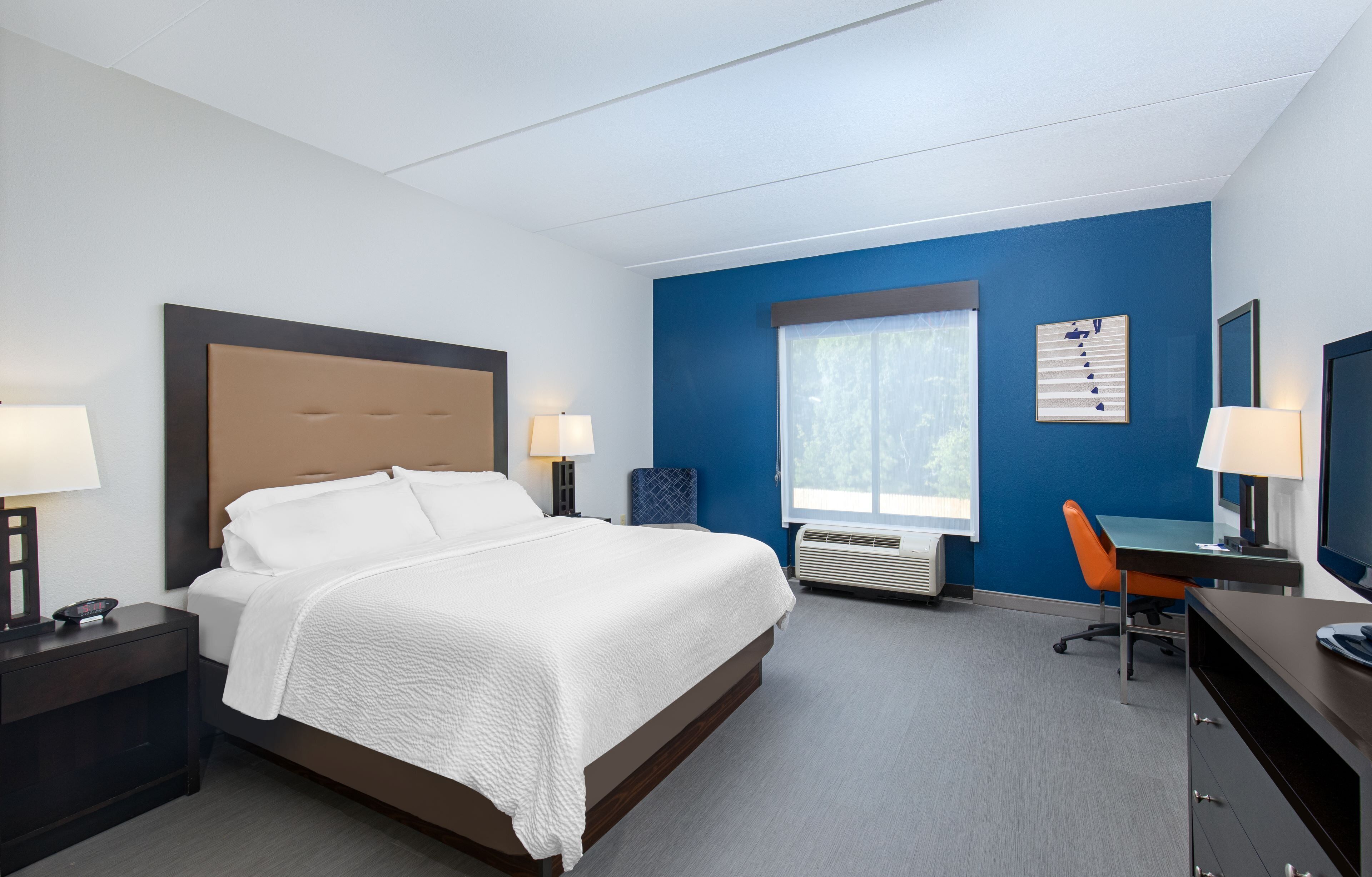 A hotel room with a bed, bedside tables, a desk, a chair, a window, and a wall-mounted TV.