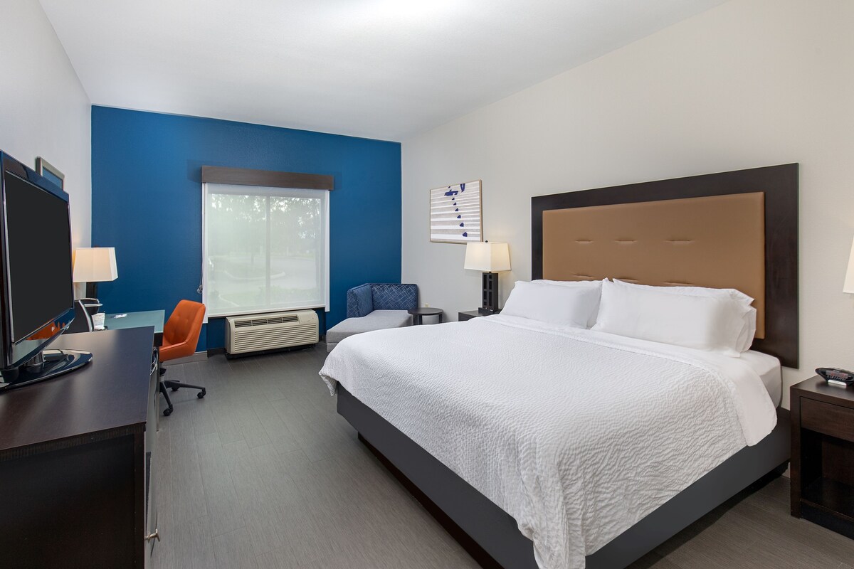 A hotel room with a large bed, a desk with a computer, a chair, a window with blinds, and a blue accent wall.