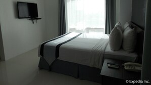 A hotel room with a bed, a TV mounted on the wall, a bedside table with a remote control, and a window with curtains. - Cityscape Hotel (Mandaue)