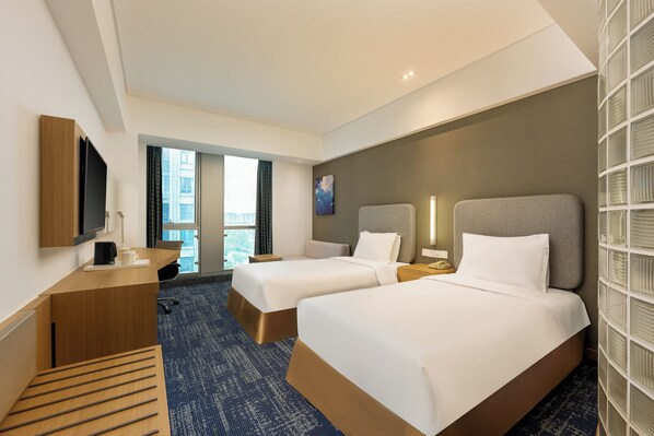 A hotel room with two beds, a desk, a TV, and a view of the city.