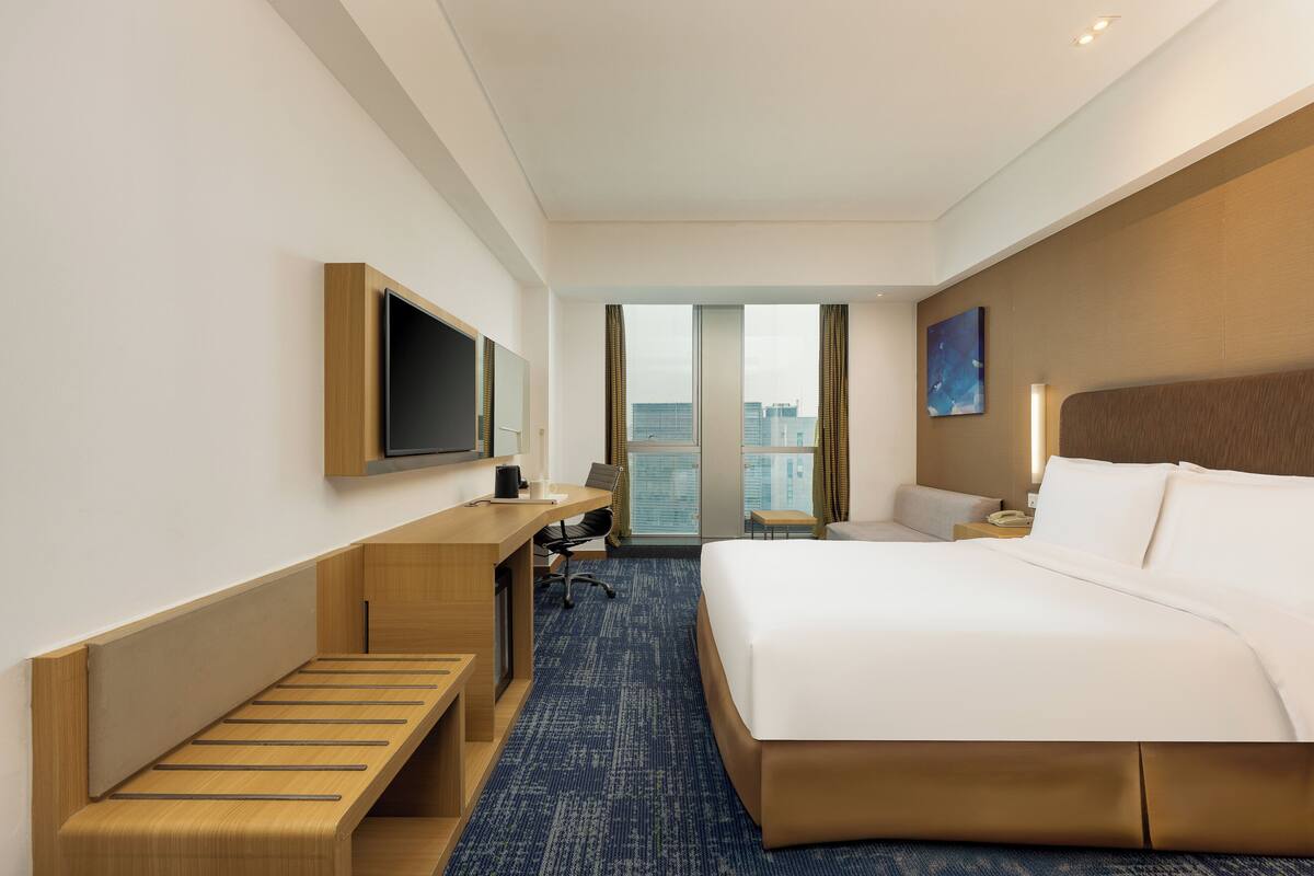 Ein Hotelzimmer mit Bett, Schreibtisch, Stuhl und einem großen Fenster mit Blick auf die Stadt.