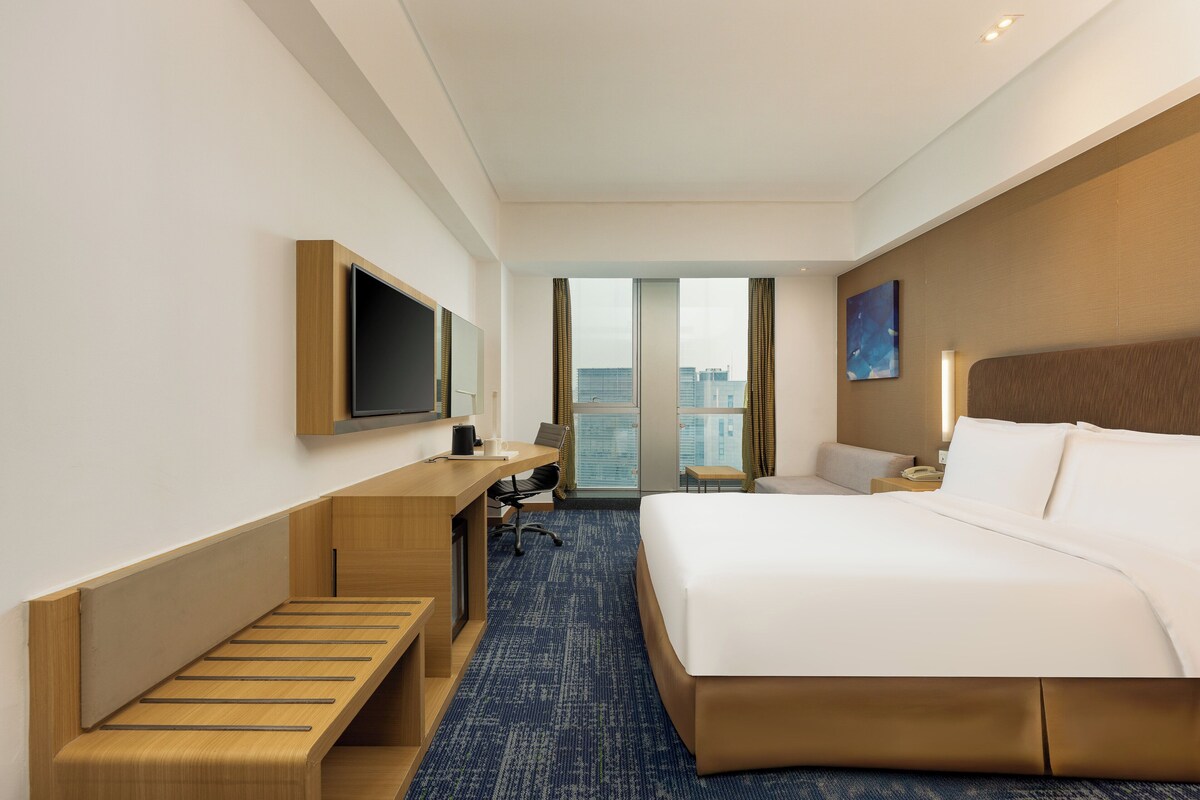 A hotel room with a bed, desk, chair, and a large window offering a city view.