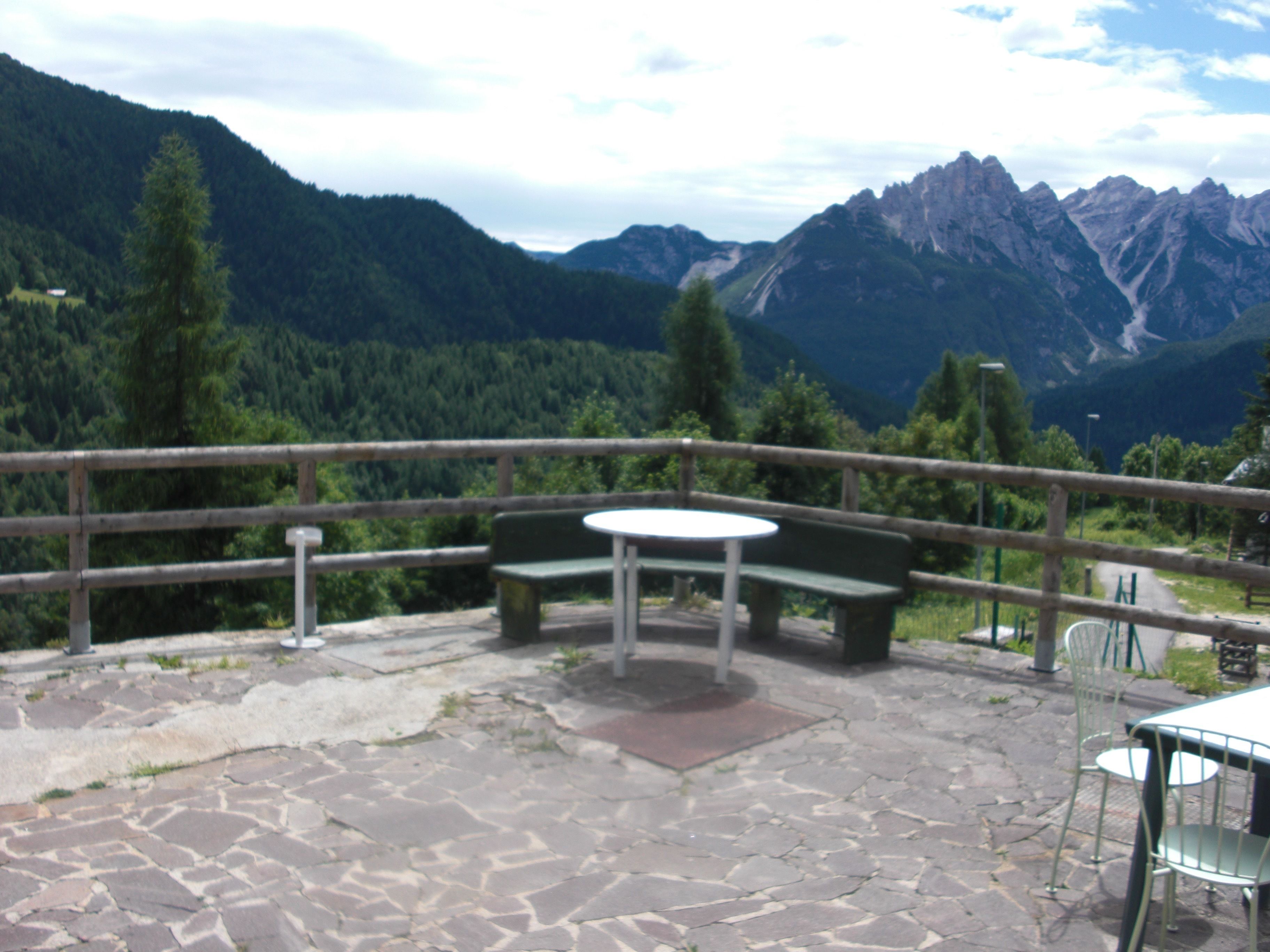 Un espace extérieur aménagé avec une table et des bancs, entouré d’une terrasse pavée de pierres et d’une rambarde en bois.