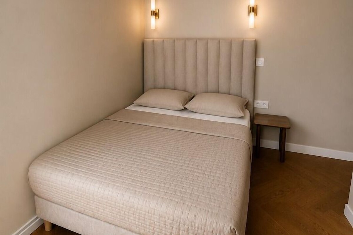 A bedroom with a large bed, two wall-mounted light fixtures, a nightstand, and a wooden floor.