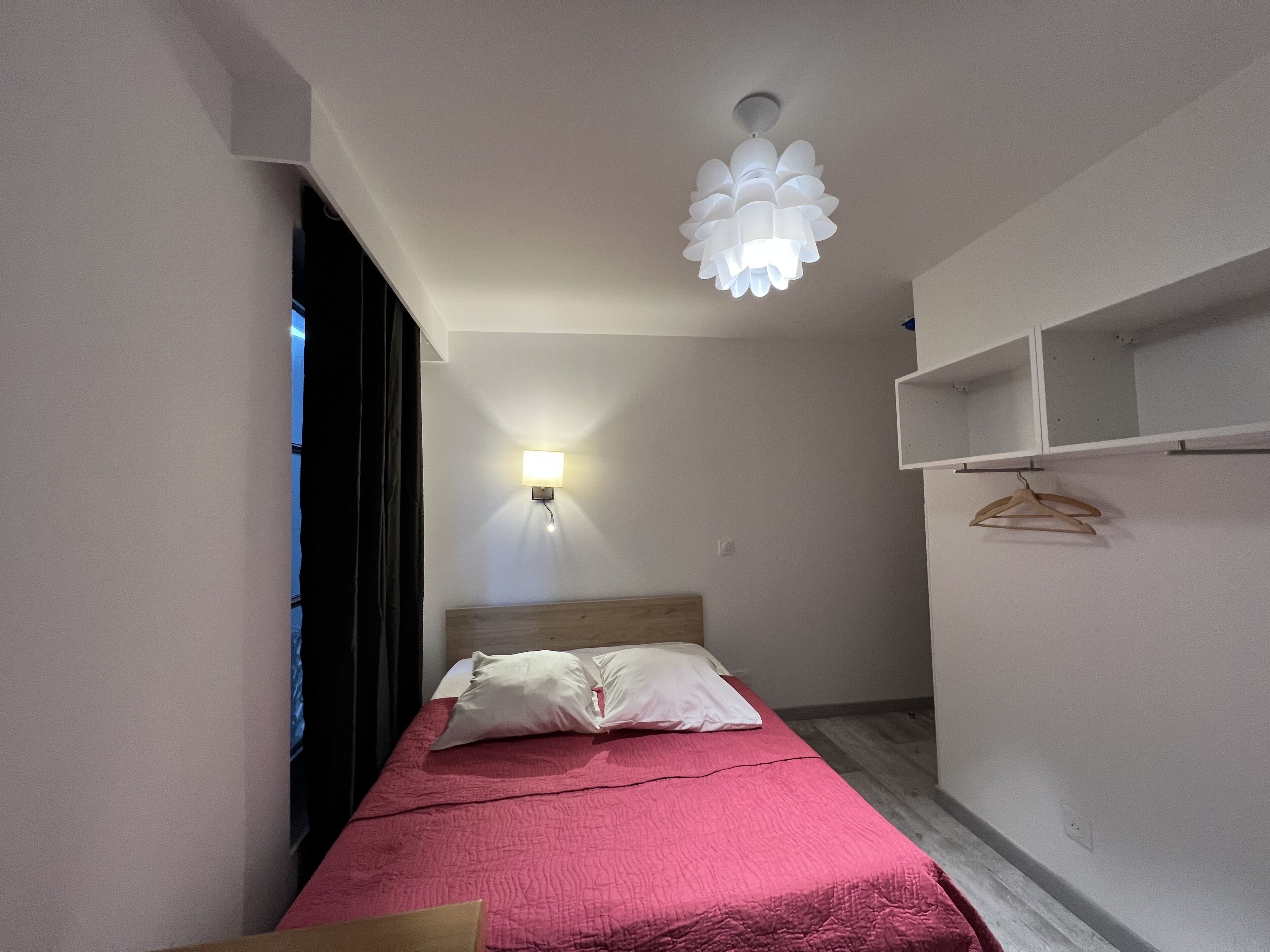 A narrow hotel room with a bed, a hanging light fixture, and a wall-mounted shelf with hangers.