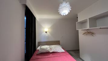 A narrow hotel room with a bed, a hanging light fixture, and a wall-mounted shelf with hangers.