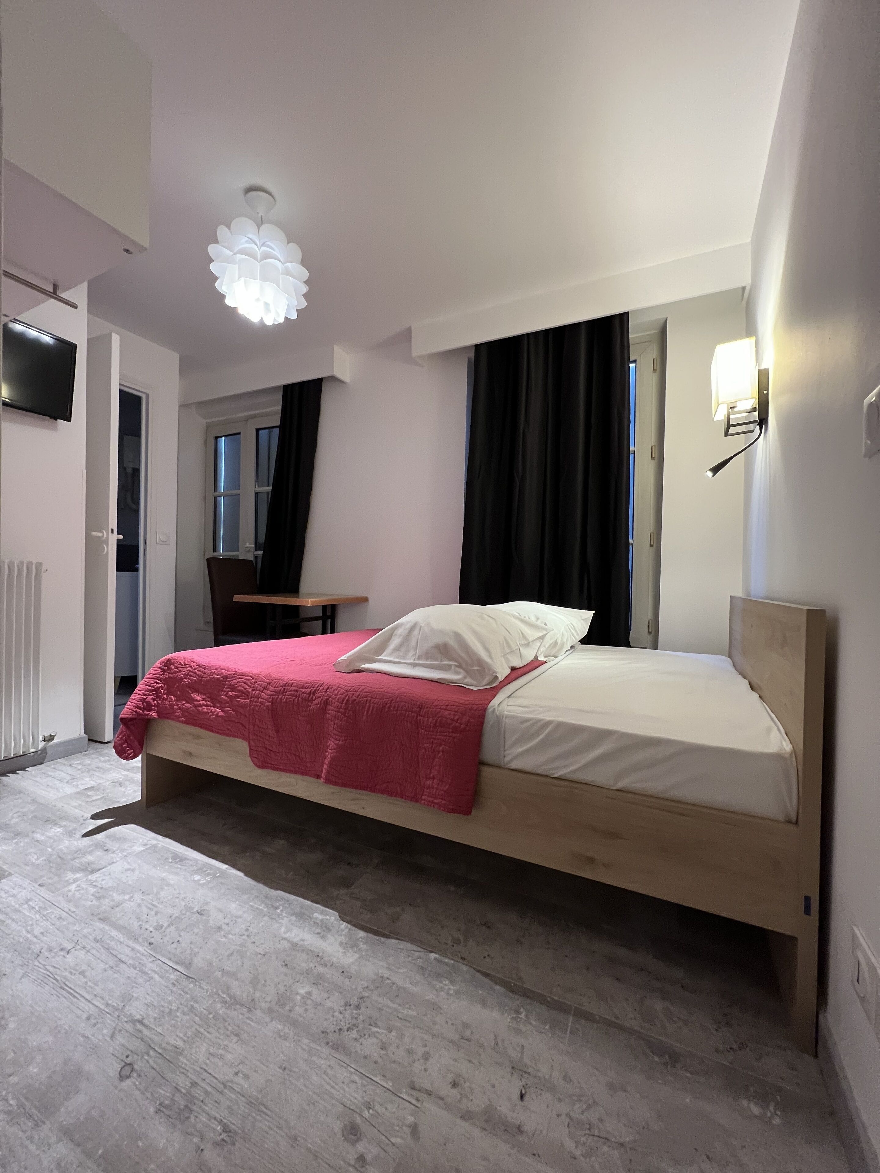 A bedroom with a bed, a desk, a chair, a TV, and a window with curtains.