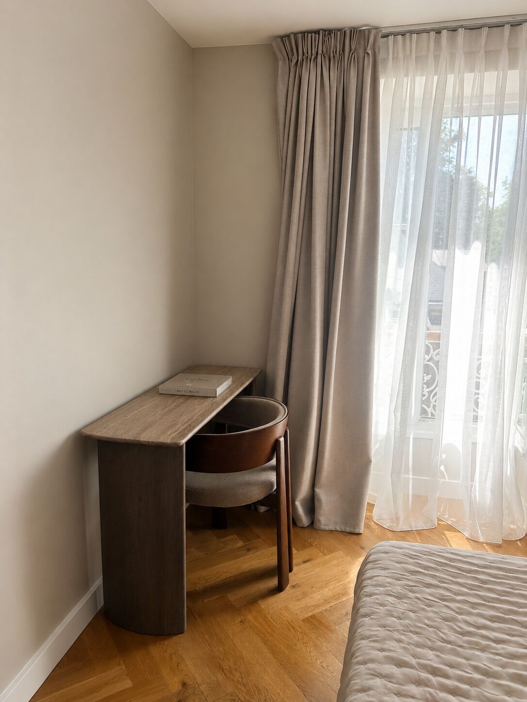 A corner bedroom with a wooden desk, a chair, a bed, and sheer curtains.