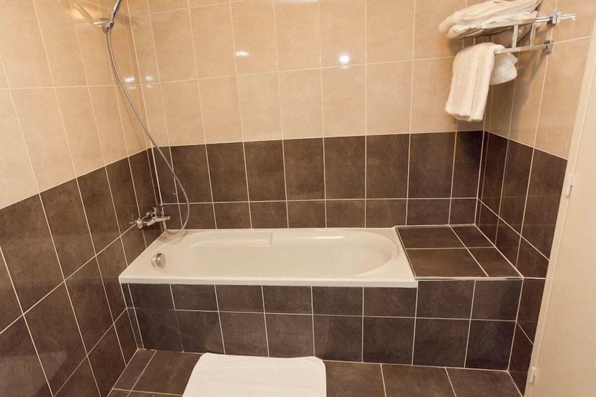 Un baño con bañera, baldosas oscuras, un toallero con toallas y un cabezal de ducha.