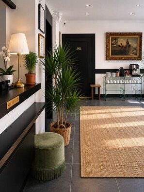 A reception area with a reception desk, a potted plant, a lamp, a green ottoman, a black door, a framed painting, and a coffee station. - Hotel Belle Fontainebleau (Fontainebleau)