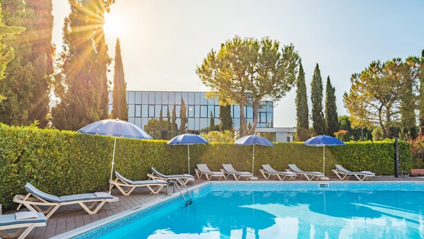 Una piscina con lettini e ombrelloni, circondata da siepi e alberi.