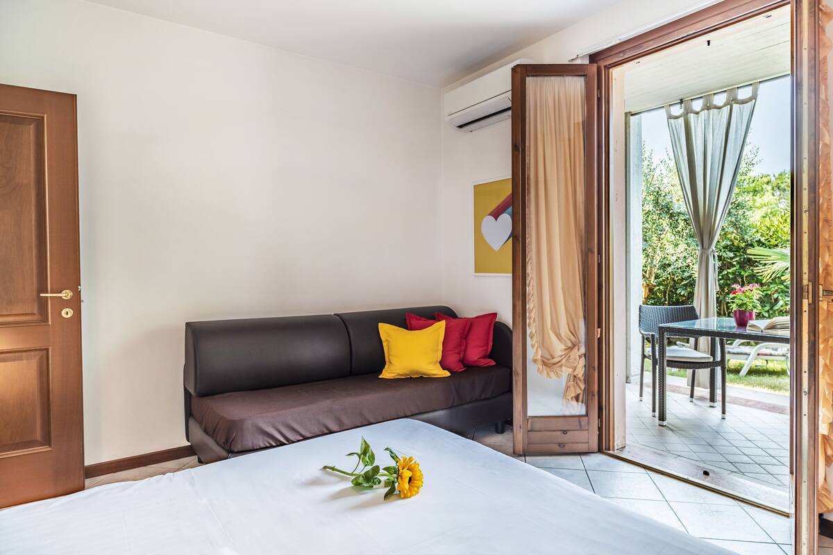 A room with a bed, a brown sofa with colorful cushions, a dining table with chairs, and a sliding glass door leading to an outdoor area.