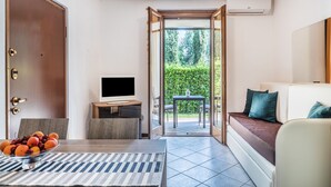 Una camera moderna con un divano, una TV su un tavolino, un tavolo da pranzo con della frutta e vista su un giardino attraverso una porta aperta.