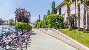 Una fila di biciclette parcheggiate lungo un marciapiede, accanto a un edificio con una barriera in cemento e vegetazione.