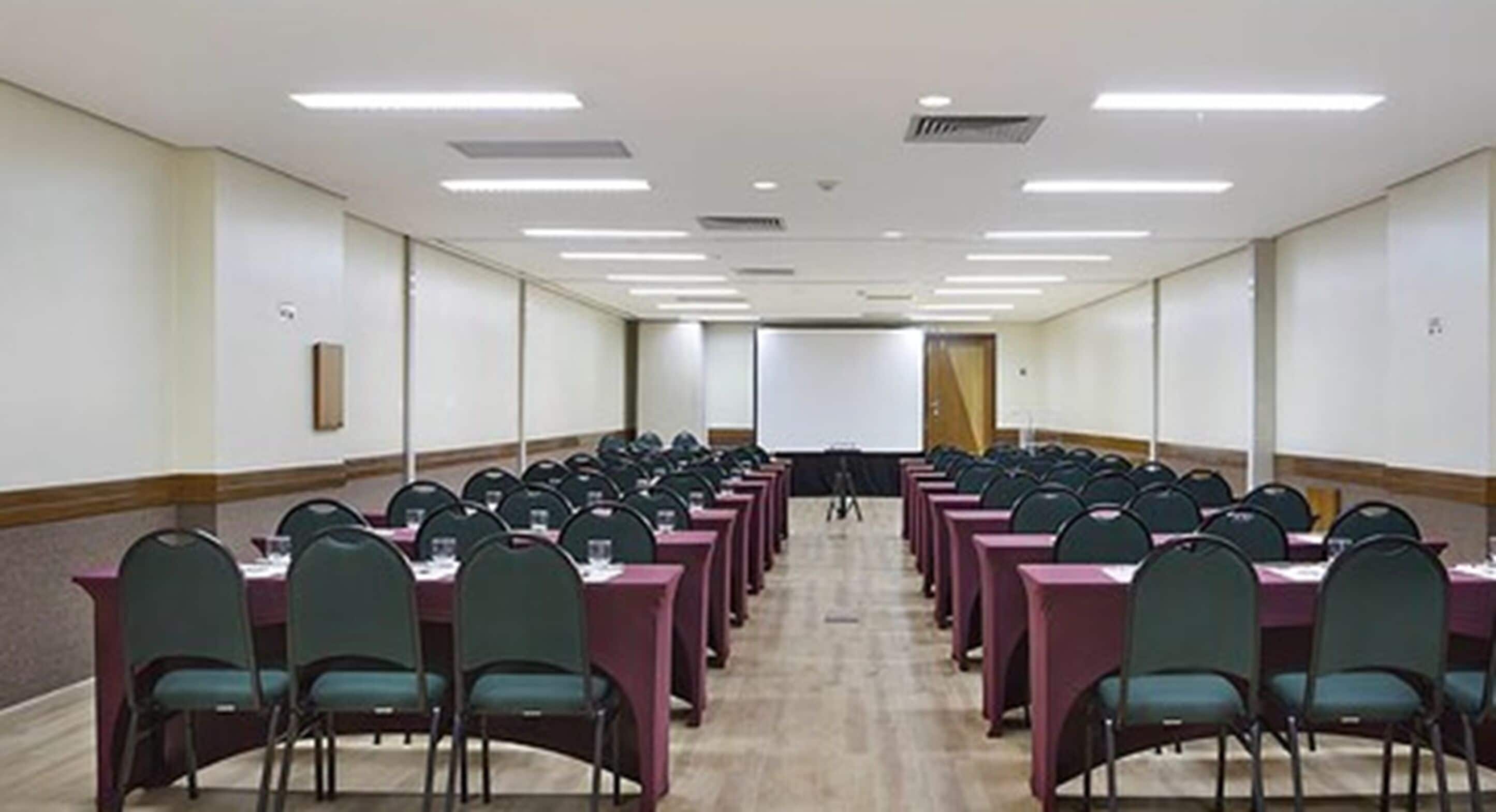 Un salón de conferencias con filas de mesas y sillas, equipado con proyectores y una pizarra.