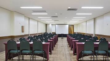 Un salón de conferencias con filas de mesas y sillas, equipado con proyectores y una pizarra.