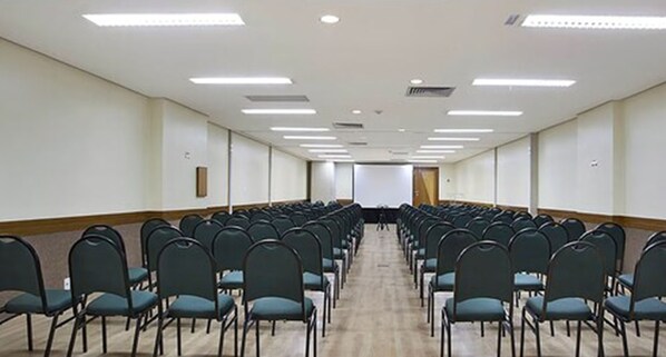 Un salón de conferencias con filas de sillas, una pantalla para proyector y luces en el techo.