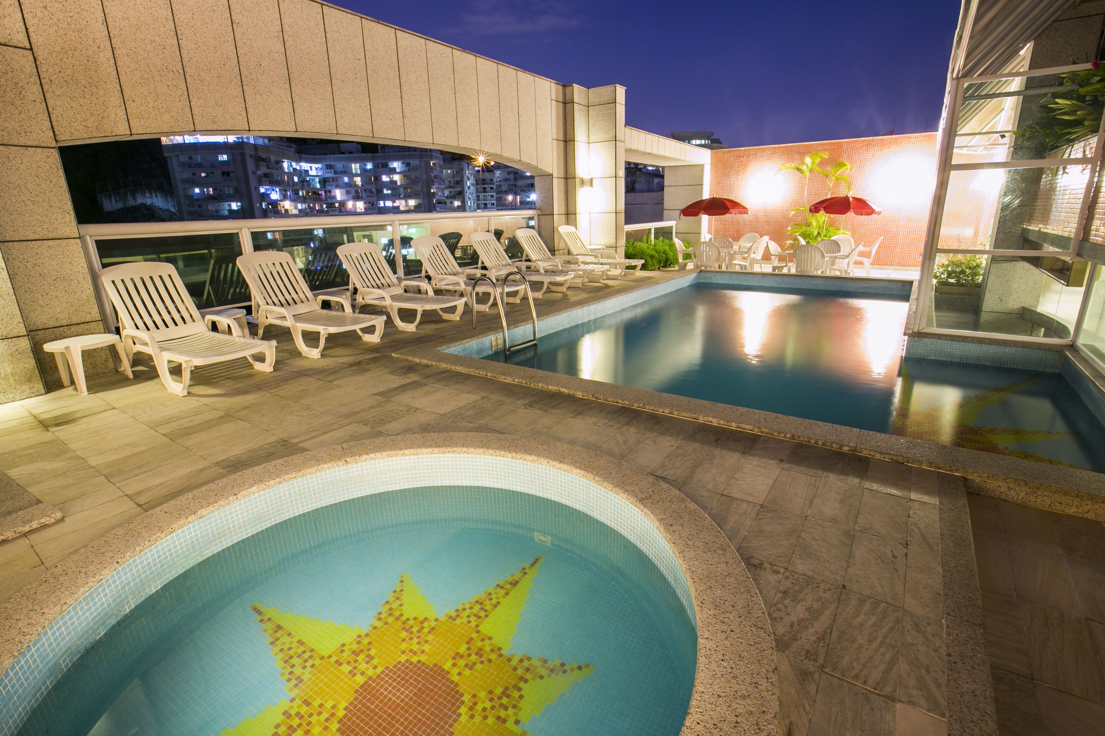 A rooftop pool area with lounge chairs and umbrellas, a decorative sun pattern in the pool, and a cityscape view at dusk.
