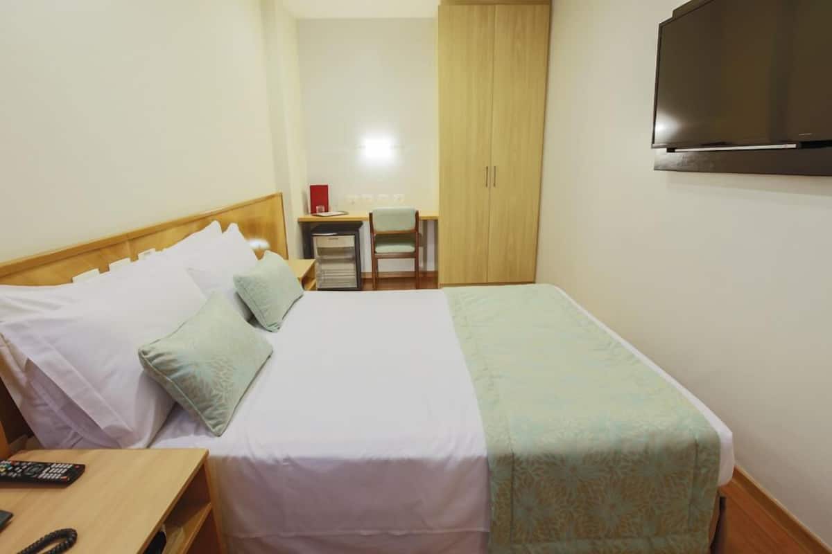 A hotel room with a bed, a desk with a chair, a wardrobe, and a TV mounted on the wall.