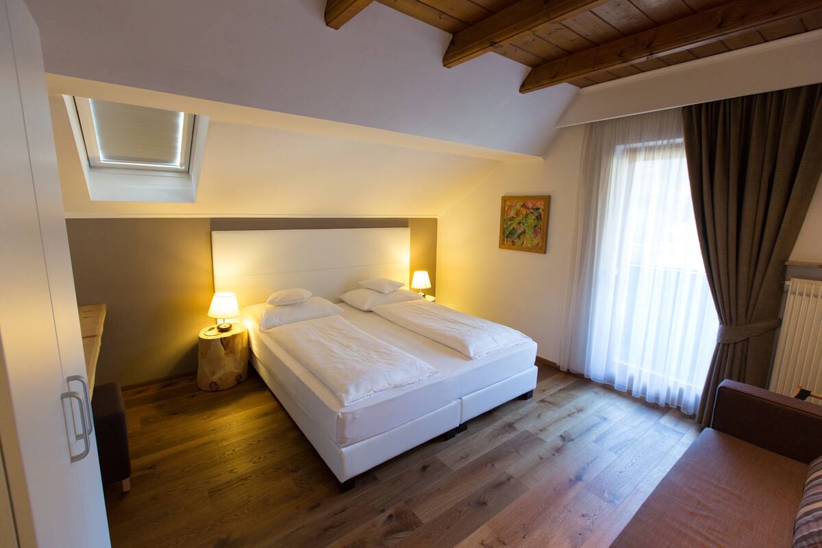 Een slaapkamer met een groot bed, dakraam, nachtkastje met lamp en raam met gordijnen.
