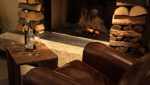 Un coin salon confortable avec une cheminée, une table avec des bouteilles et des verres, et une pile de bois de chauffage.