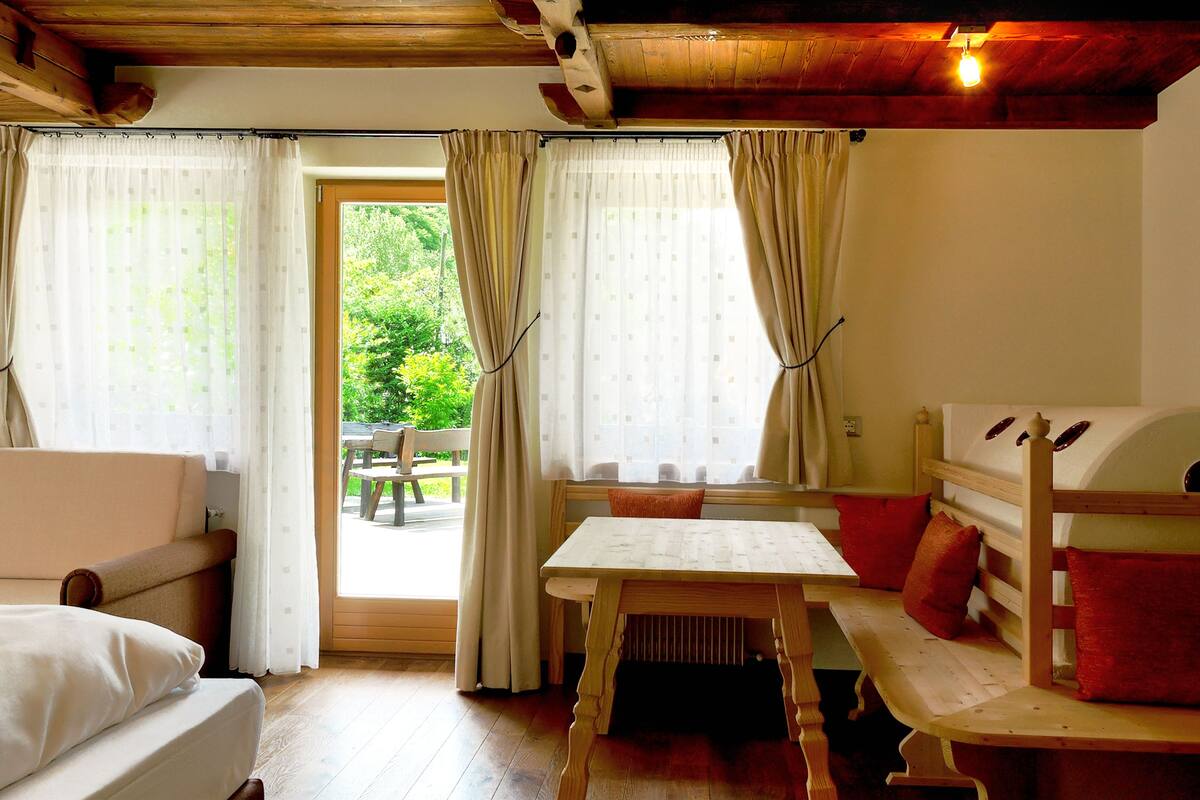 Een knus vertrek met een bed, een tafel en een bankje. Er is een raam met gordijnen en uitzicht op groen.