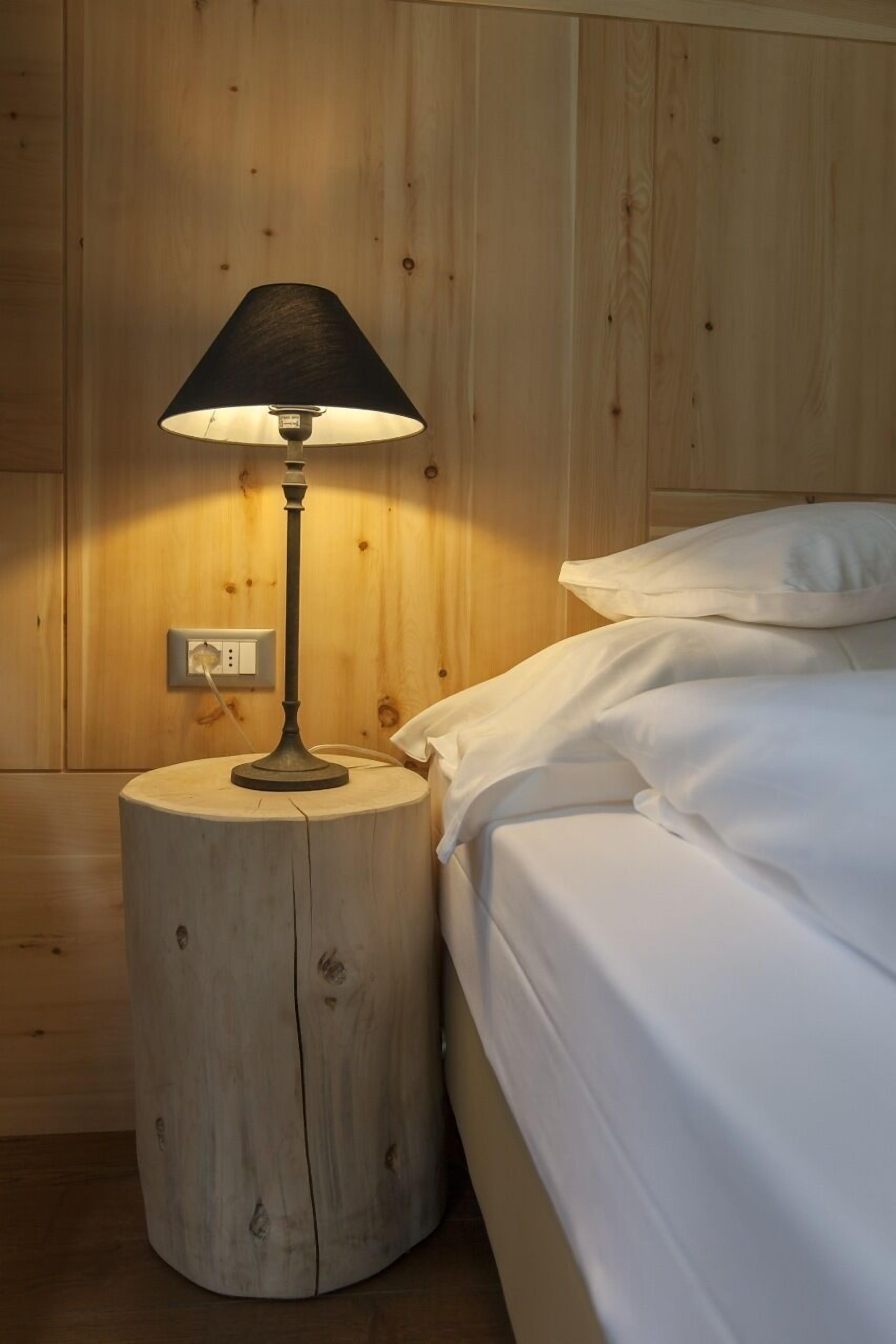 Ett träbord som står vid sängen, med en lampa på, en säng med vita sängkläder och ett vägguttag.