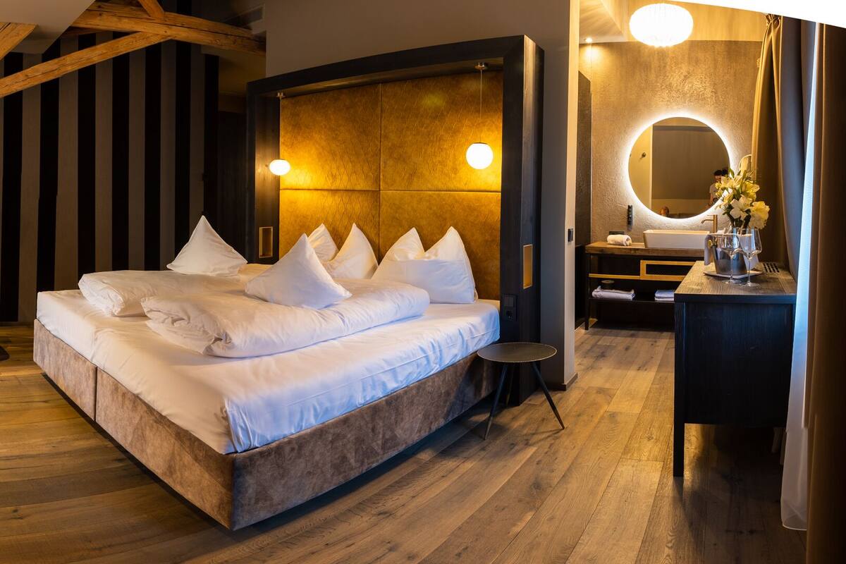 Een moderne hotelkamer met een groot bed, een zithoek met een fauteuil en een klein tafeltje, een kaptafel met spiegel, en een houten plafondconstructie.