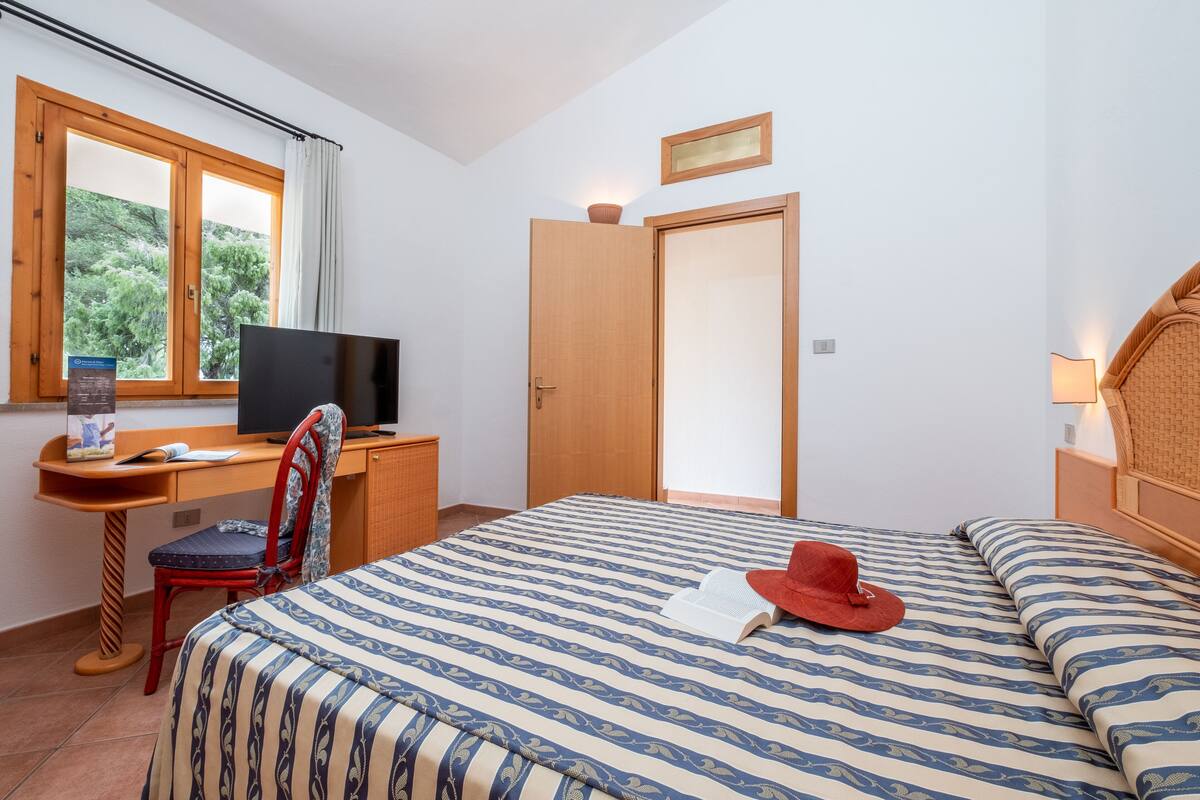 A bedroom with a bed, a desk with a computer, a chair, a television, and a door.