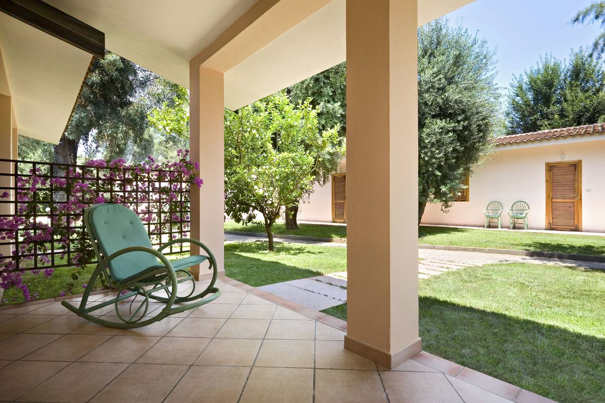 A covered patio area with a rocking chair, a small garden, and a building with a tiled roof in the background.