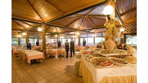 A spacious indoor venue with high ceilings and wooden beams, featuring multiple tables set for dining, a buffet area with various food items, and a view of the outdoors through large windows. - TH Marina di Sibari - Baia degli Achei Village (Cassano allo Ionio)