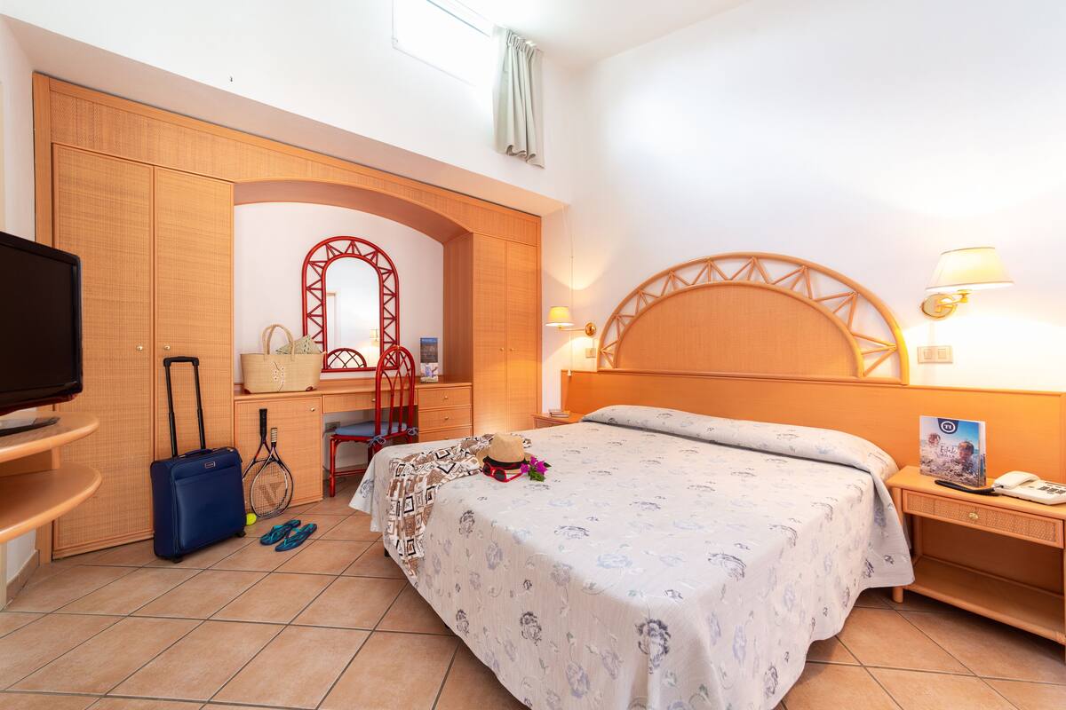A hotel room with a bed, a desk, a chair, a TV, a suitcase, and a tennis racket.