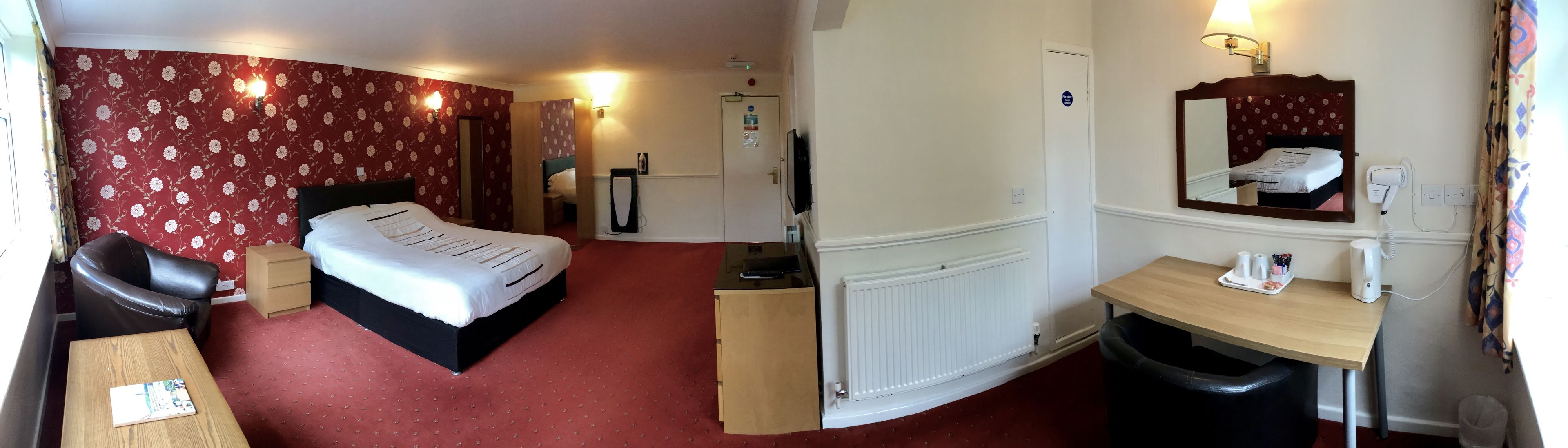A hotel room with a bed, a desk, a chair, a radiator, a mirror, and a trash bin.