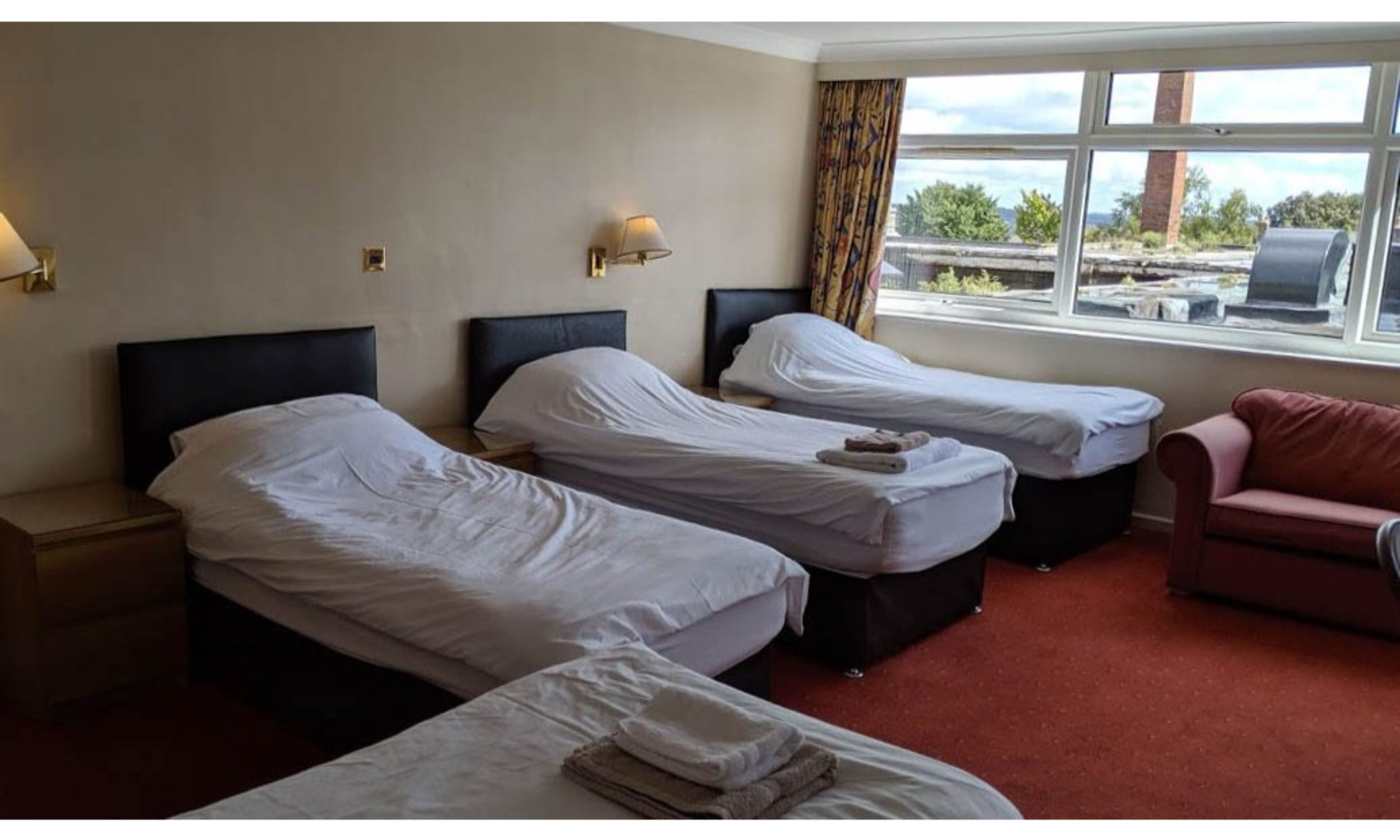 Ein Hotelzimmer mit zwei Einzelbetten, einem roten Schlafsofa und einem Fenster mit Blick nach draußen.