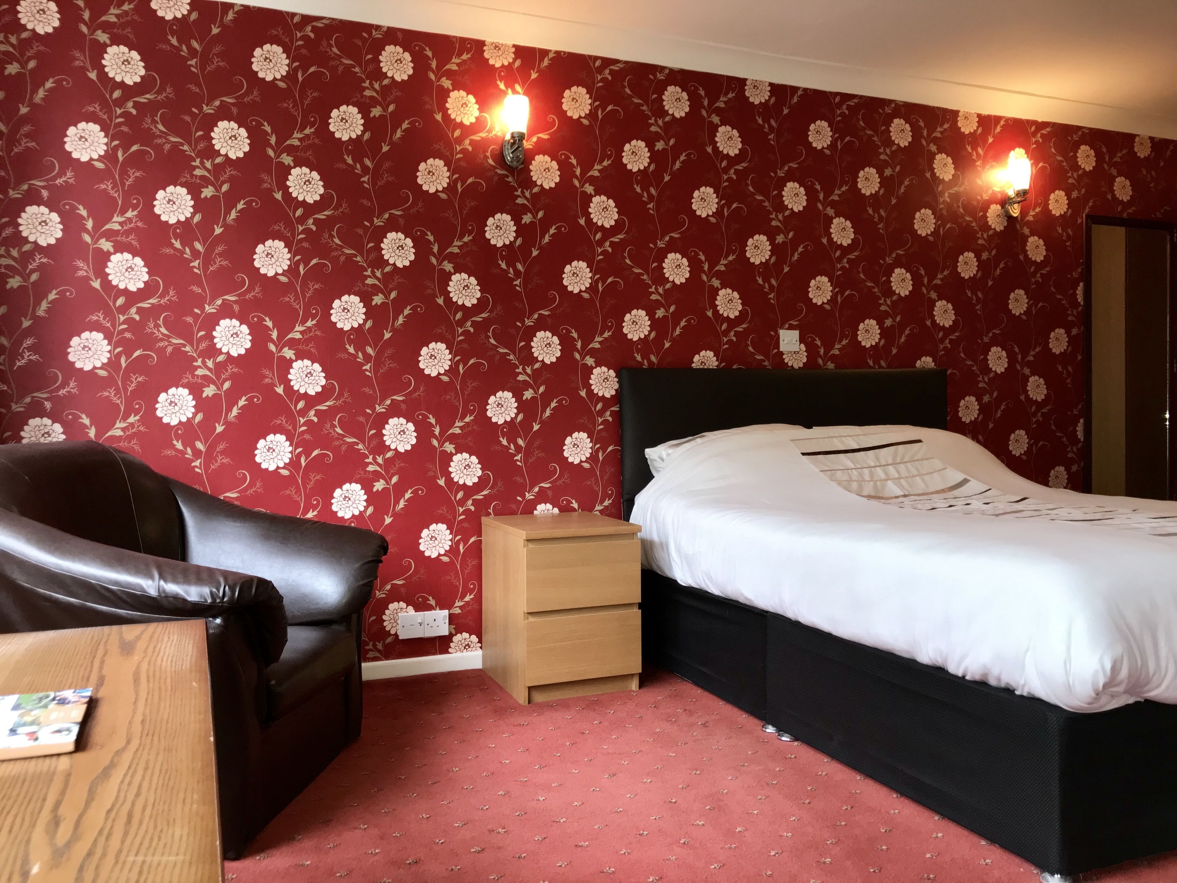 A bedroom with a floral wallpaper, a black headboard, a wooden nightstand, a black leather chair, and a red carpet.