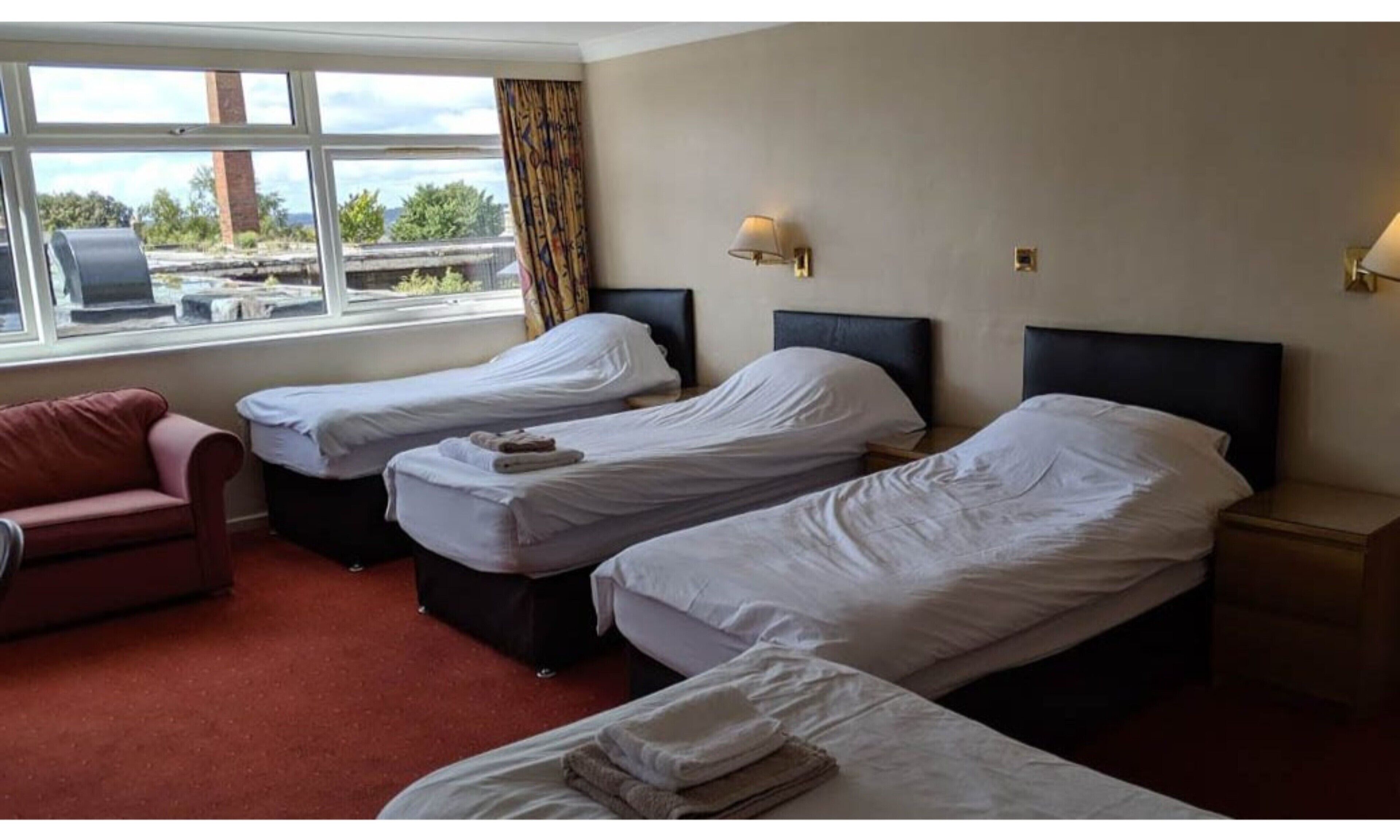 A hotel room with two single beds, a red sofa, a window with a view of the outside, and a lamp on the wall.