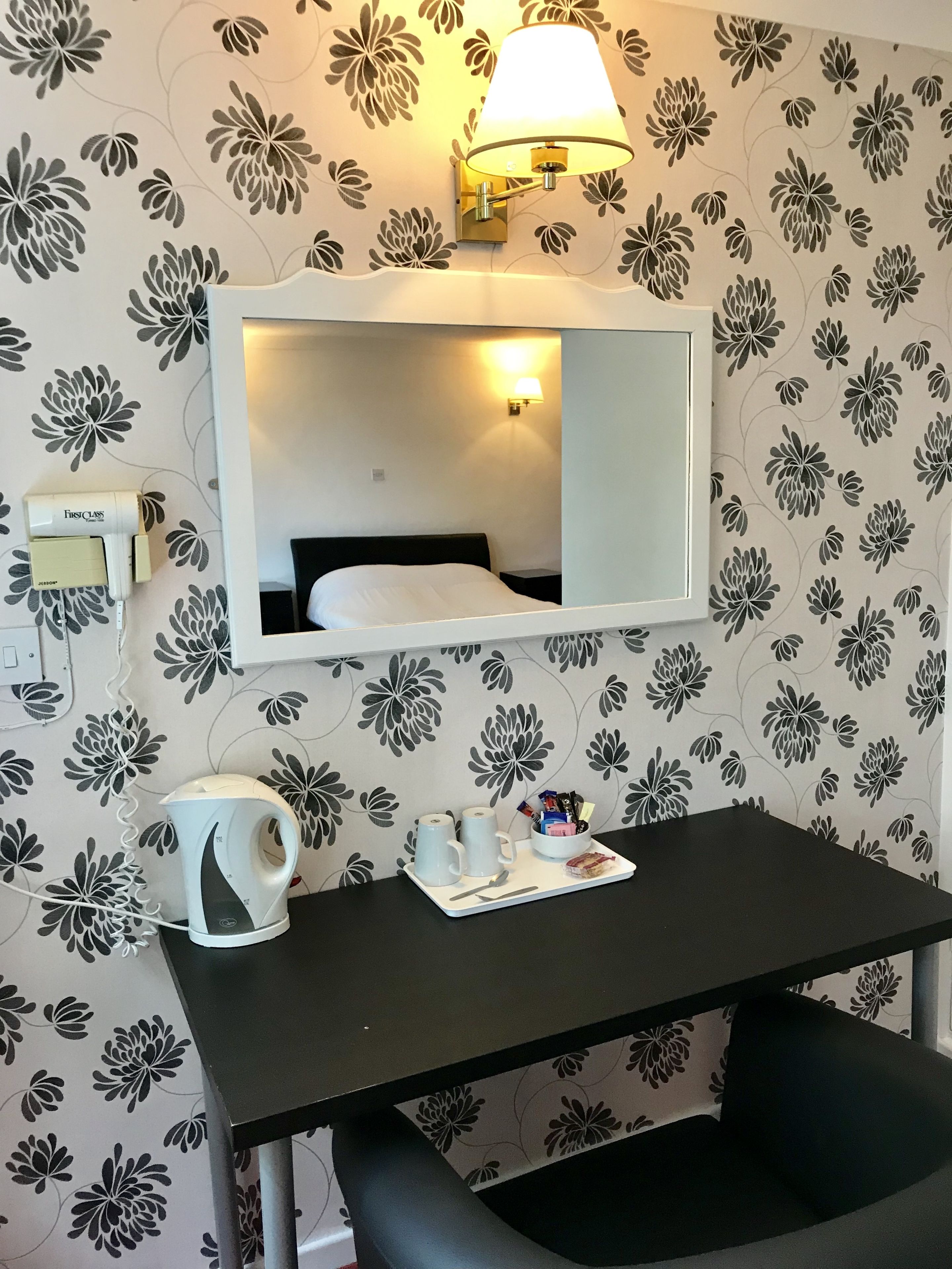 A hotel room with a bed, a desk with a kettle and mugs, a lamp, and a FirstClass hairdryer.