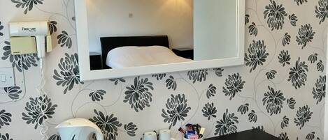 A hotel room with a bed, a desk with a kettle and mugs, a lamp, and a FirstClass hairdryer.
