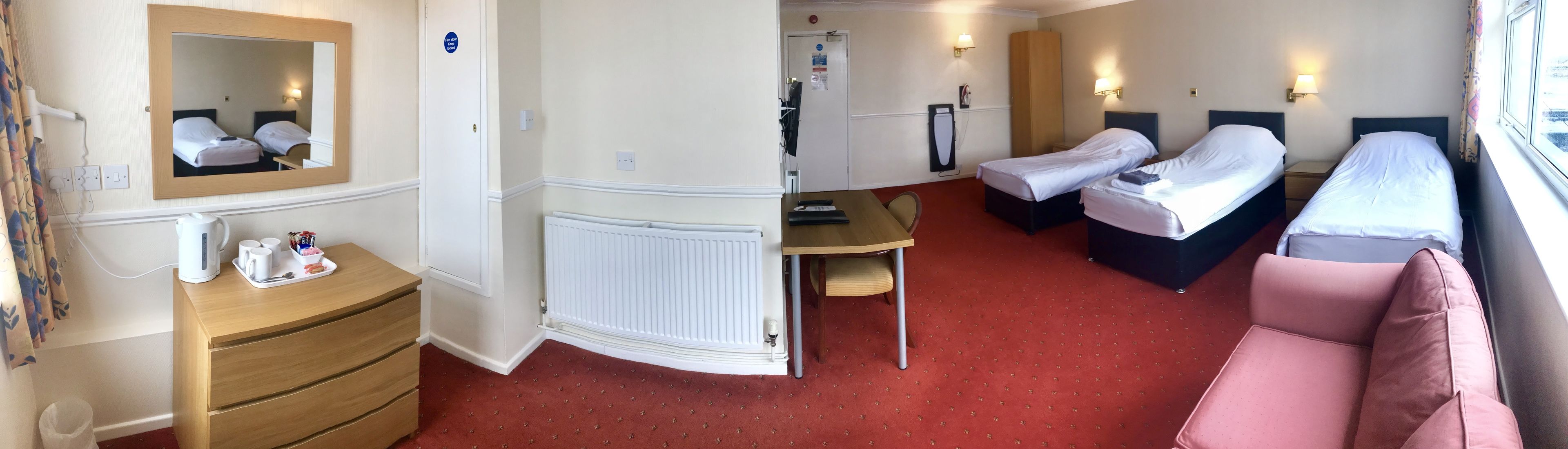 A hotel room with two beds, a wooden dresser, a radiator, a small table with a chair, and a window with curtains.