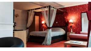 Ein Himmelsbett mit roten Blütenmuster-Tapete, ein Nachttisch mit Lampe und ein roter Sessel.