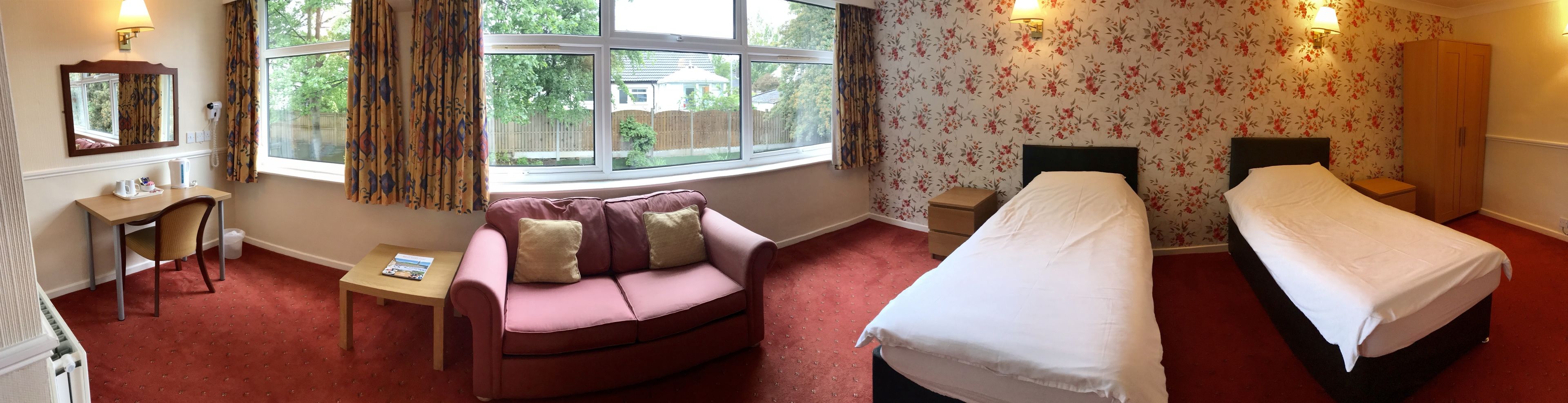 A hotel room with a sofa, two single beds, a small table, a mirror, and a window with curtains.