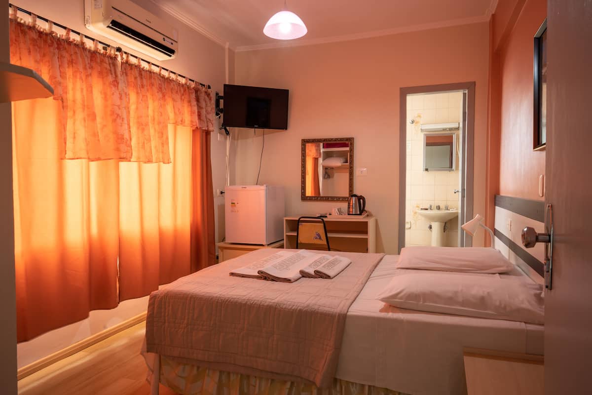 Habitación de hotel con cama, escritorio, televisor, aire acondicionado y baño visible a través de una puerta abierta.