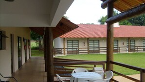 Un área de estar al aire libre con techado, mesas y sillas, una valla de madera y un edificio con techo rojo al fondo.