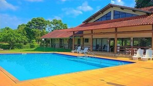Una piscina con borde de baldosas, una zona de estar techada con sillas y un edificio con techo de tejas rojas.
