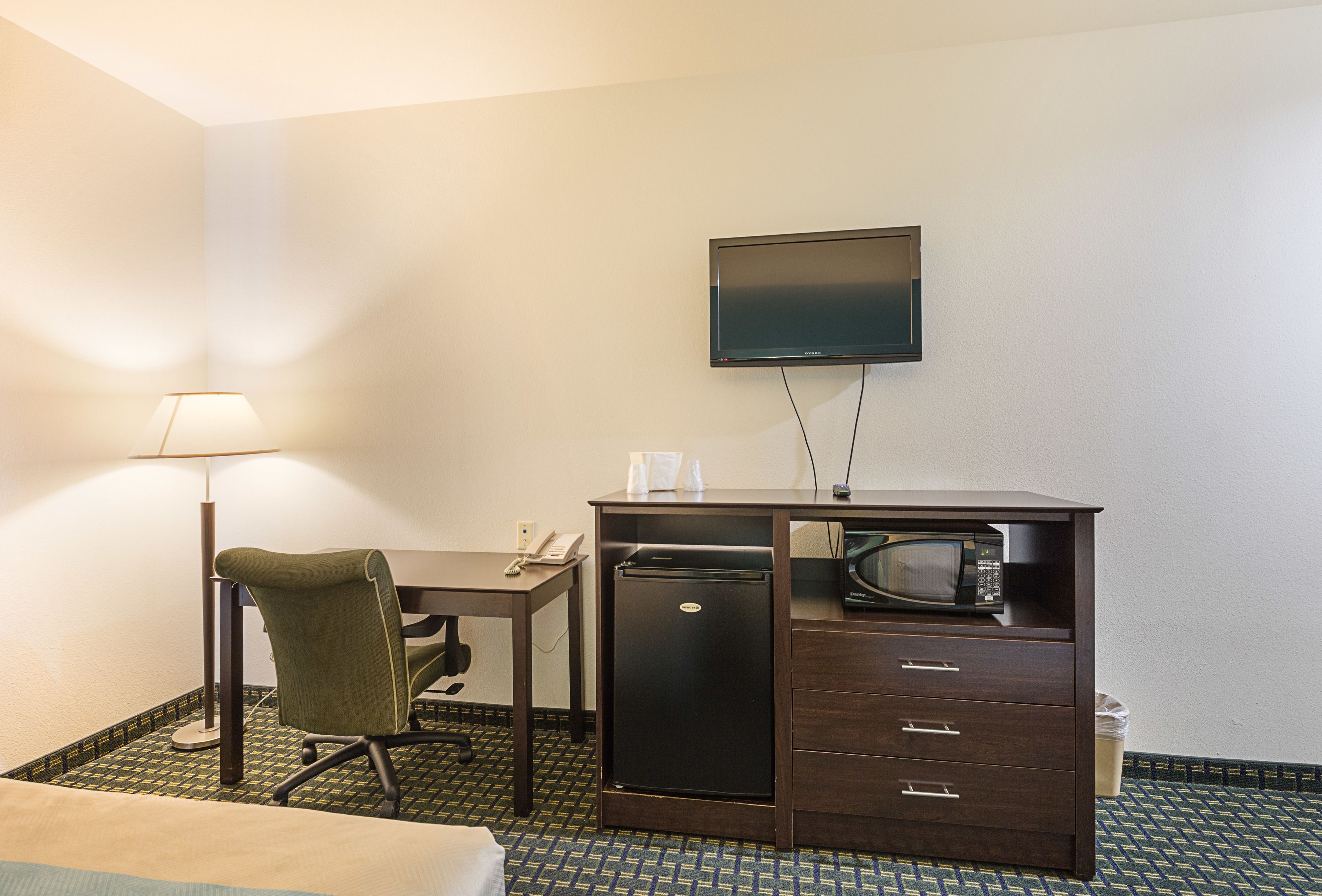 A hotel room with a desk, chair, flat-screen TV, refrigerator, microwave, and a lamp.
